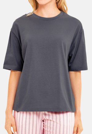 Frau, die ein locker sitzendes dunkelgraues T-Shirt mit kurzen Ärmeln und rosa-weiß gestreifte Hose trägt, gezeigt von den Schultern bis zur Hüfte.