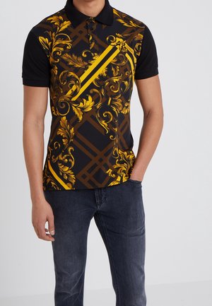 Mann trägt ein schwarzes Poloshirt mit gelben und braunen barocken Blumen- und geometrischen Mustern, kombiniert mit dunkelblauen Jeans.