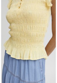 Top amarillon con fruncido en los hombros, que presenta una tela texturizada y un dobladillo con volantes. Llevado sobre una falda de mezclilla azul.