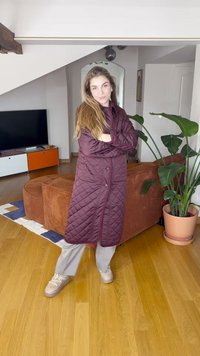 Ilgas siuvinėtas paltas bordo spalvos, su segėjimo mygtuku ir rombų raštu. Dėvimas ant šviesiai pilkų kelnių ir smėlio spalvos avalynės. Vidaus aplinka.