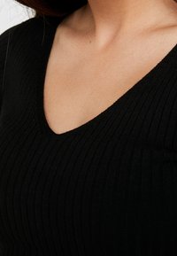 Primo piano di una persona che indossa una maglietta nera a coste con scollatura a V, che mostra il collo e la parte superiore del petto.
