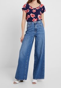 Hvězdný top s květinovým potiskem v námořnické modři s růžovými květy, kombinovaný s vysokým pasem a širokými modrými džíny. Doplňují ho bílé, stylové sandály na otevřenou špičku.