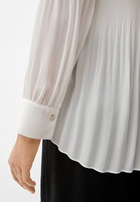 Blouse blanche fluide avec tissu froncé, manches longues et poignet lisse avec un détail de bouton métallique. Texture douce et légère.