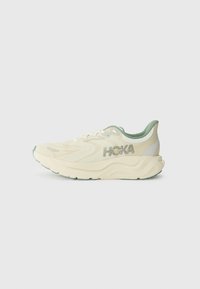 Hoka-Laufschuh mit einem strukturierten cremefarbenen Obermaterial, grünen Akzenten, dicker weißer Zwischensohle und einer schwarzen Gummisohle, die ein Logo an der Seite aufweist.