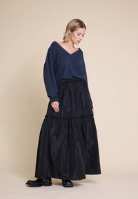 Marineblå strikkegenser med V-hals og avslappede ermer, kombinert med et svart, lagdelt maxiskjørt laget av blankt stoff med elastisk midjebånd.