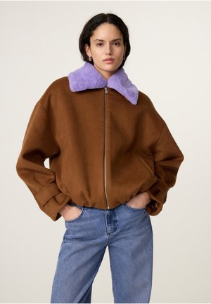 Woman wearing a brown zip-up jacket with a large purple fur collar and blue jeans, standing with hands in pockets against a plain background.