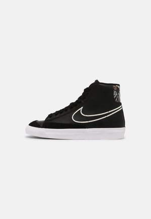 Czarny sneaker za kostkę z białą podeszwą i dużym białym logo Nike, z wzorzystym detalem na kołnierzu przy pięcie.