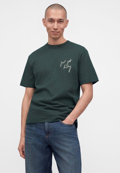 Jeune homme à la tête rasée portant un t-shirt vert foncé avec le texte "just get lucky" et un jean bleu, debout avec une main dans la poche.