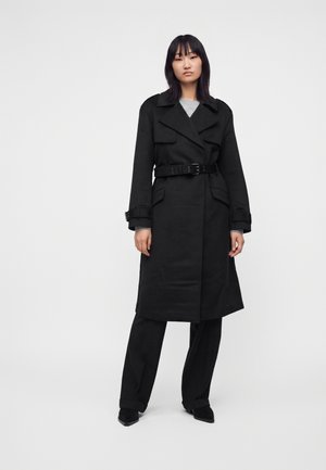 Femme portant un long trench-coat noir ceinturé sur un pantalon et des chaussures noirs, debout devant un fond clair uni.