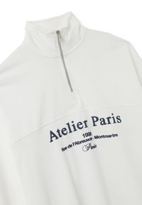 Pullover bianco con colletto alto e zip a quarti. Presenta testo ricamato blu navy con la scritta "Atelier Paris" e ulteriori dettagli sottostanti.