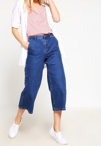 Kvinde iført løse blå denimjeans, en lyserød mønstrets top, en hvid åben skjorte og hvide slip-on sko, stående med den ene hånd i lommen.