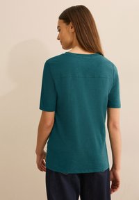 Camiseta de manga corta en color azul verdoso, hecha de una tela ligera, que presenta un corte relajado y un escote ligeramente redondeado con un detalle de costura en la parte posterior.