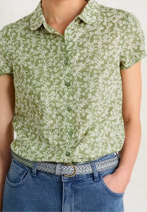 Frau trägt ein grünes, kurzärmeliges Hemd mit Blumenmuster, das in blaue Jeans gesteckt ist, mit einem hellgrauen geflochtenen Gürtel und einer Hand in der Hosentasche.