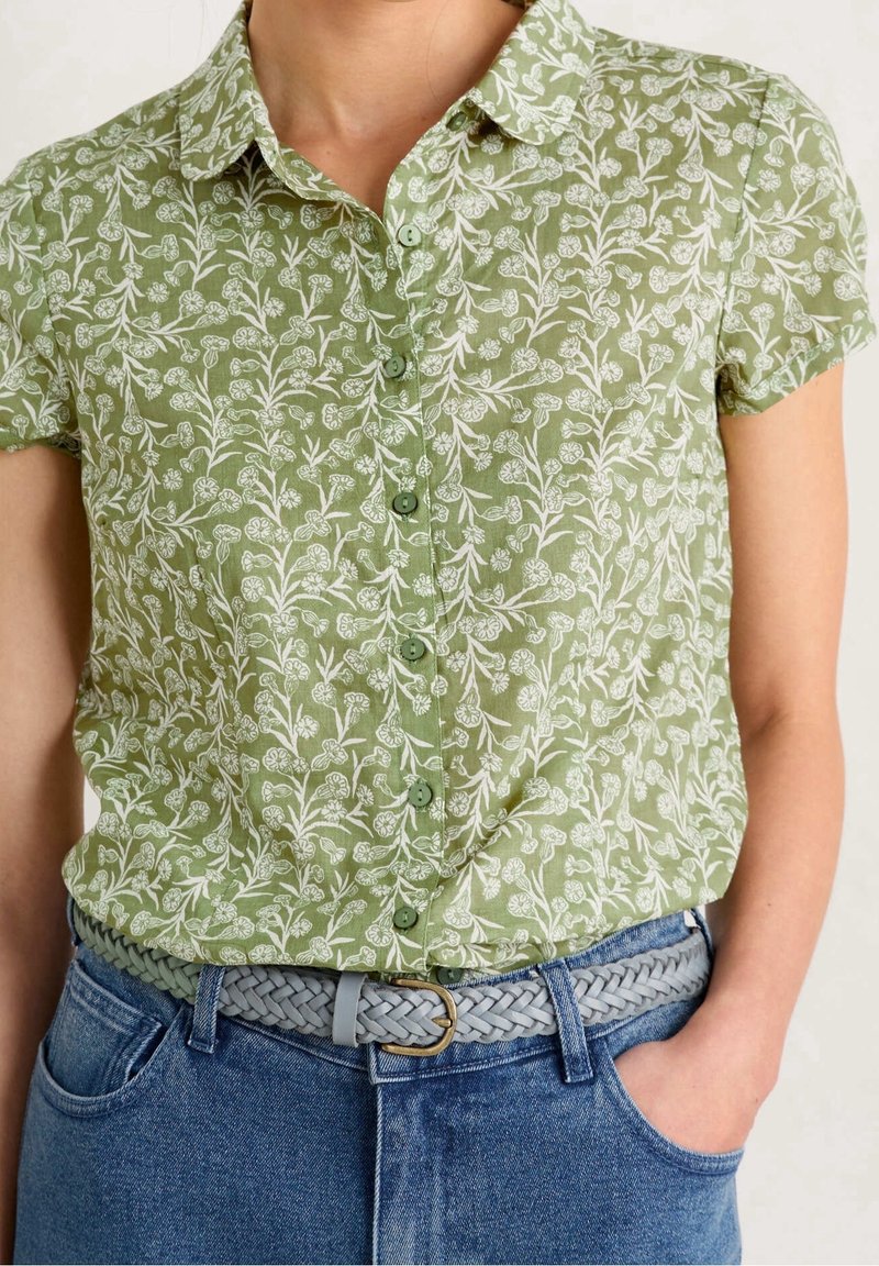 Femme portant une chemise verte à manches courtes à fleurs boutonnée, rentrée dans un jean bleu avec une ceinture tressée gris clair et une main dans la poche.