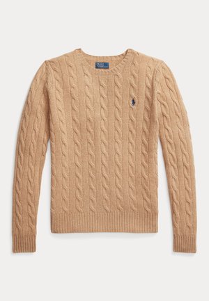 Kvinne som står med hendene i lommene, iført en beige kabelstrikket genser, blå jeans og brune mokasinsko mot en ensfarget bakgrunn.