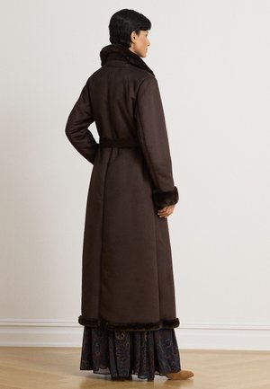 Femme debout de dos, portant un long manteau marron foncé bordé de fourrure sur une jupe à motifs et des chaussures beige, devant un mur uni.