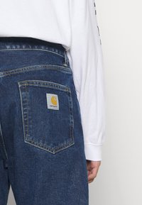 Vaqueros de mezclilla con un lavado azul oscuro, que presentan un bolsillo trasero cuadrado con un parche del logo de Carhartt en amarillo y blanco y detalles de costura visibles.