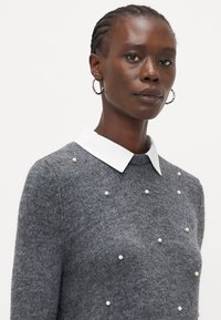 Grå genser med rund hals, hvit krage og dekorative perler ordnet på fronten. Strikket tekstur med slim fit design.
