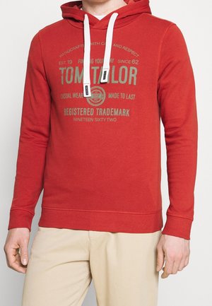 Uomo che indossa una felpa con cappuccio rossa Tom Tailor con coulisse bianche e pantaloni beige, in piedi davanti a uno sfondo chiaro e semplice.