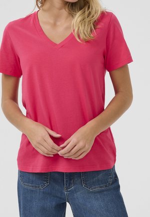 Mujer que lleva una camiseta rosa brillante de manga corta con cuello en V y vaqueros azules de mezclilla, de pie con las manos entrelazadas al frente.