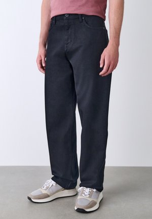 Mann trägt lockere schwarze Hose, beige und weiße Sneaker und ein rosa-rotes Hemd, steht auf grauem Boden vor einer weißen Wand.