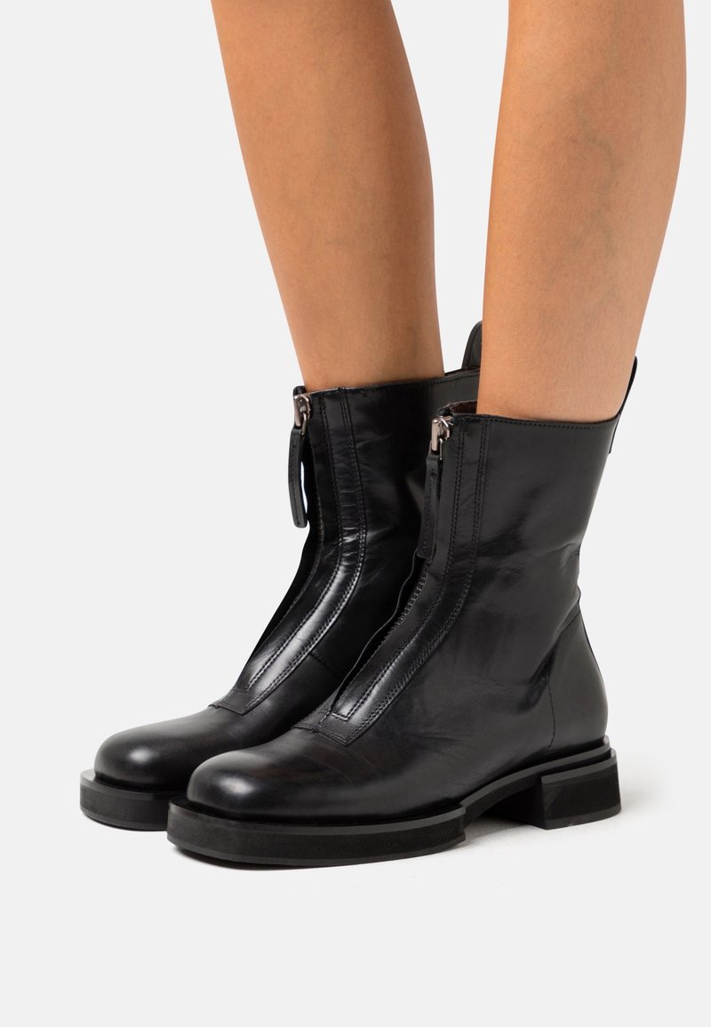 Bottes noires en cuir mi-mollet avec fermeture éclair à l'avant et semelle épaisse, portées par une personne, vues de profil sur un fond blanc.