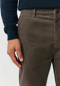 Die linke Hand einer Person ruht in der Nähe der Tasche einer braunen Cordhose und trägt ein dunkelblaues Langarmshirt.