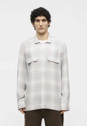 Άνδρας με σγουρά μαλλιά που φοράει ένα ανοιχτό γκρι καρό πουκάμισο με τσέπες με καπάκια, στέκεται μπροστά σε ένα απλό λευκό φόντο.