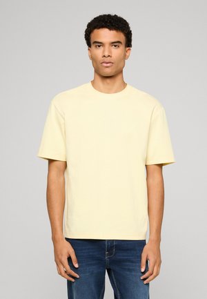 Young man with curly hair wearing a plain light yellow short-sleeve T-shirt and blue jeans, standing against a gray background.