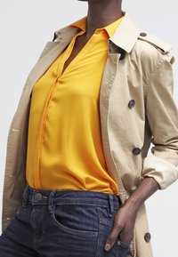 Beige trenčkot přes jasně žlutou blůzu s véčkovým výstřihem na knoflíky, spárovaný s tmavými džínami s zakřiveným designem kapes.