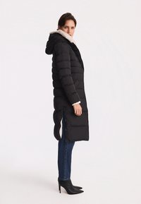 Czarna kurtka puchowa z kapturem i białym futrem, z zamkiem błyskawicznym i bocznymi rozcięciami, noszona na niebieskich dżinsach i czarnych szpilkach.