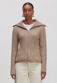 Beige Strickjacke mit Reißverschluss, mit geripptem Muster, hohem Kragen und langen Ärmeln. Getragen über einem weißen T-Shirt, kombiniert mit hellen Hosen.