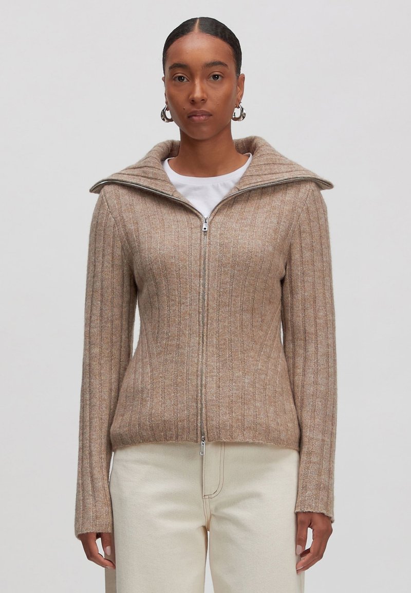 Beige Strickjacke mit Reißverschluss, mit geripptem Muster, hohem Kragen und langen Ärmeln. Getragen über einem weißen T-Shirt, kombiniert mit hellen Hosen.