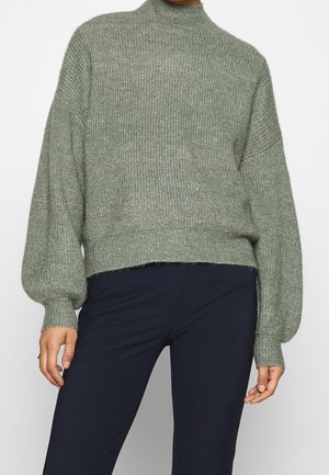 Mujer con suéter verde de punto acanalado con mangas globo y cuello alto, combinado con pantalones oscuros de corte ajustado.