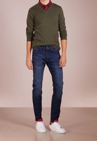 Ung man i olivgrön tröja över mörk vinröd skjorta, mörkblåjeans, röda strumpor och vita sneakers står på beige golv.