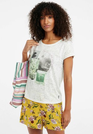 T-shirt grigio chiaro con un motivo testurizzato e un design grafico multicolore, abbinato a pantaloni corti floreali gialli e una borsa a tracolla a righe.