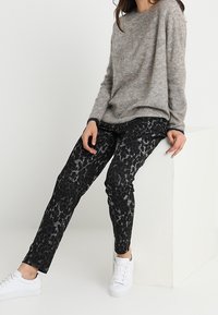Grå strikketrøje med løs pasform, parret med sorte mønstrede skinny bukser. Hvide sneakers fuldender outfittet. Minimalistisk baggrund.