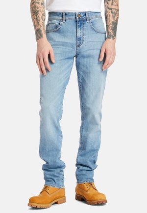 Jasnoniebieskie jeansy o dopasowanym kroju noszone z beżowymi, roboczymi butami, ukazujące tatuaże na ramionach i dłoniach po bokach, na jednolitym tle.