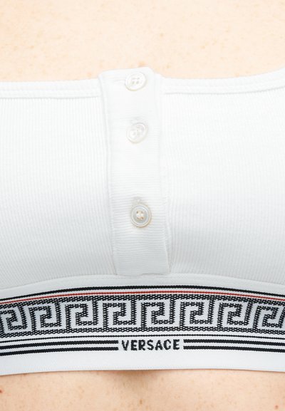 Κοντινό πλάνο λευκού κορσάζ με ριγέ υφή, τρία κουμπιά και μαύρη ελαστική ταινία με μοτίβο, η οποία φέρει την επιγραφή «Versace», σε φόντο δέρματος.