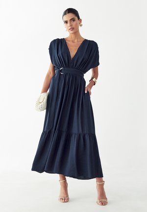 Kobieta w granatowej, maxi sukience z paskiem i głębokim dekoltem w serek, trzymająca białą plecioną kopertówkę, nosząca złotą biżuterię i beżowe szpilki.