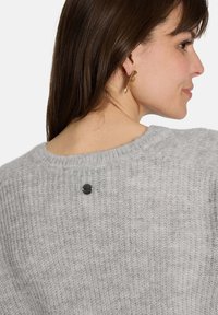 Grauer Strickpullover mit weicher Textur, Rundhalsausschnitt und einem schwarzen Knopfdetail im Rücken. Goldfarbene, blattförmige Ohrringe sichtbar.