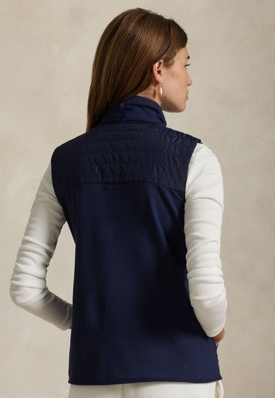 Γιλέκο σε ναυτικό μπλε με επενδεδυμένο πάνω μέρος και ομαλή κάτω επιφάνεια. Γιακάς που στέκεται όρθιος, λογότυπο στην πλάτη και εφαρμοστό σχέδιο για κομψή εμφάνιση.