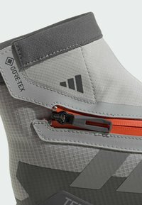 Gray Gore-Tex boot featuring a textured upper, orange zipper, and black accents. The design includes a rounded shape and a cushioned collar.