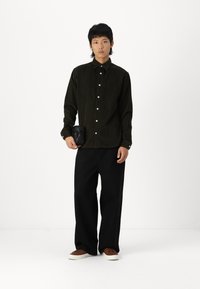 Mørkegrønn kordfløyels skjorte med knapper, overdimensjonerte svarte bukser, brune joggesko og en liten svart crossbody veske. Enkel bakgrunn.