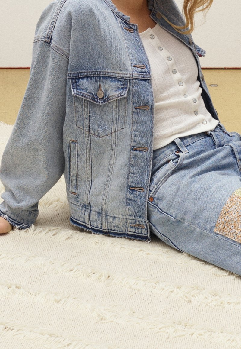 Světle modrá džínová bunda s volným střihem, opatřená dvěma náprsními kapsami a hnědými knoflíky, kombinovaná s bílou košilí na knoflíky a džínovými kalhotami.