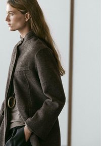Brown wool-blend coat with a stand collar, front pocket, and loose fit. Worn over a gray top, with a large black pendant accessory.
