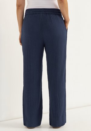 Persoon draagt losse, marineblauwe textuurbroek en een wit mouwloos topje, staand met handen in de zakken tegen een effen achtergrond.