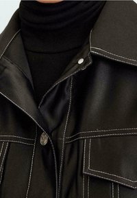 Zwarte satijnen jack met witte stiksels, een puntige kraag en een drukknoop. Het ontwerp omvat voorzakken en een gladde textuur.