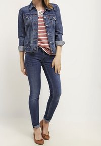 Denim jasje met lange mouwen, knoopsluiting, twee borstzakken, gedragen over een gestreept rood en wit top. Strakke blauwe jeans en bruine schoenen.