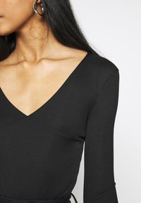 Kvinna iklädd en svart ribbad V-ringad långärmad topp och svarta och vita marmorerade hängande örhängen med rakt mörkt hår.